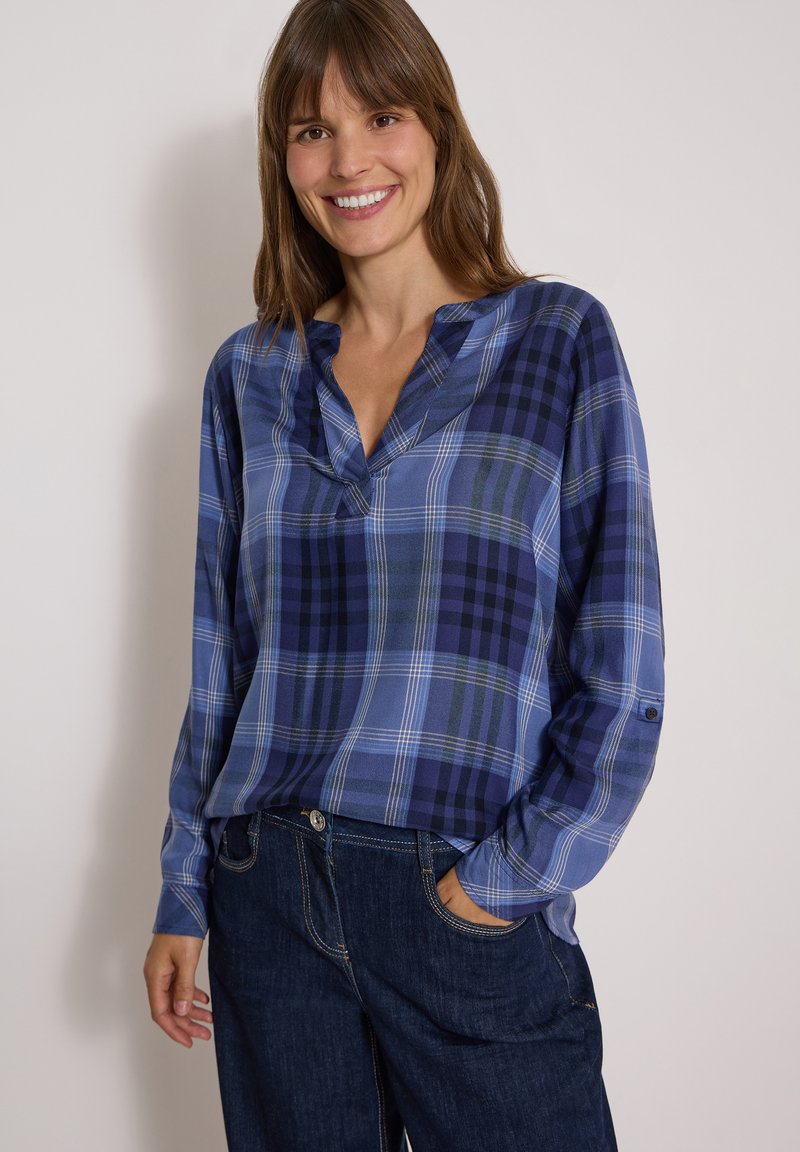 Femme souriante, portant une chemise à carreaux bleue à manches longues et un jean bleu foncé, une main dans la poche, debout devant un fond clair uni.