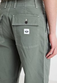 Green cargo pants in lightweight fabric with visible stitching, multiple pockets, and a branded tag on the back depicting an emblem.