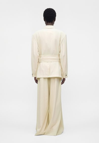 Άτομο με κοντά κατσαρά μαλλιά φοράει φαρδύ εκρού κοστούμι με σακάκι και ζώνη, στέκεται με την πλάτη προς απλό λευκό φόντο.