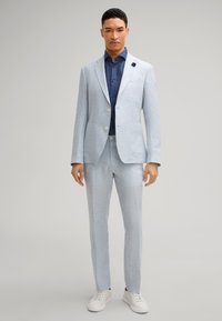 Jasnoniebieski garnitur z lnu o dopasowanym kroju, jednorzędowa marynarka, dwie przednie kieszenie, połączony z ciemnoniebieską koszulą i białymi trampkami.