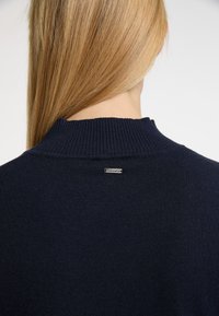 Námorní svetr s pleteným límcem, na zadní straně s malou kovovou značkou. Hladká, jemná textura a přiléhavý design.