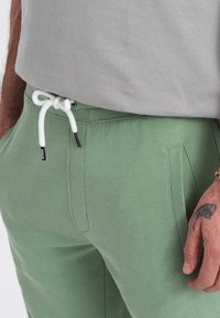 Jogging en coton vert avec ceinture côtelée, cordon blanc et poches latérales ; porté avec un t-shirt gris.