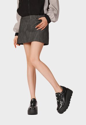 Donna con gonna corta scura, giacca bianca e nera e scarpe nere con plateau e lacci con suola spessa, in piedi con una gamba incrociata.