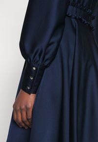 Main à la peau foncée reposant sur une robe en satin bleu marine avec manche bouffante froncée et poignet à trois boutons argentés.