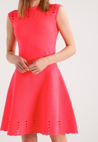 Robe sans manches ajustée de couleur rose vif avec une texture côtelée, présentant un motif découpé décoratif au niveau du col et une jupe évasée.