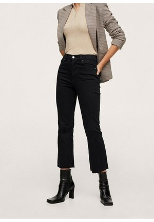 Frau trägt ein beige geripptes Oberteil, ein kariertes Sakko, schwarze, gekürzte Jeans mit ausgefransten Säumen und schwarze, hochhackige Stiefeletten, steht mit den Händen in den Taschen.