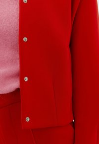 Rote Jacke mit silbernen Druckknöpfen, getragen über einem rosa flauschigen Pullover, kombiniert mit passenden roten Hosen, dargestellt vom Mittelrücken bis zum Oberschenkel.