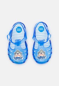 Zapatos de agua transparentes de color azul con correas, que presentan un gráfico de tiburón y el texto "¡JUGUEMOS!" en la plantilla, texturizados para proporcionar agarre y comodidad.
