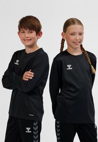 Camisetas deportivas negras de manga larga con el logo de Hummel, combinadas con pantalones negros que presentan un patrón de chevrones. Tejido suave, corte relajado.