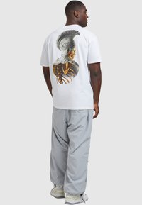 Valge T-särk, millel on graafiline trükk, mis kujutab marmorist büstiga koos kuldsete elementidega seljaosas. Hallid, laiad püksid ja kerge jalats.