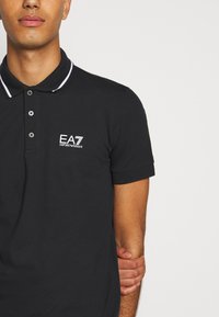 Černé polo tričko vyrobené z bavlny, s klasickým límcem s kontrastními bílými pruhy, zapínáním na dva knoflíky a vyšitým logem na hrudi.