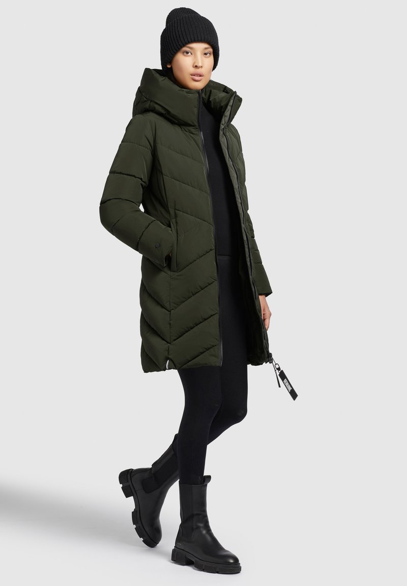 Daunenjacke in Dunkelgrün mit Kapuze, präsentiert ein Chevron-Steppdesign, mit Reißverschluss und seitlichen Taschen. Kombiniert mit schwarzen Leggings und Stiefeln.