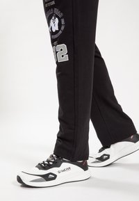 Melni sporta bikses ar grafisku apdruku sānos, kombinētas ar baltiem un melniem sporta apaviem ar teksturētu augšdaļu un saites.