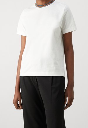 Personne portant un T-shirt blanc à manches courtes avec un col rayé multicolore et un pantalon noir ample, debout devant un fond uni.