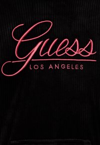Musta samettikangas, jossa näkyvä vaaleanpunainen kirjailtu "Guess"-logo, ja sitä alhaalla pienemmällä tekstillä "LOS ANGELES".