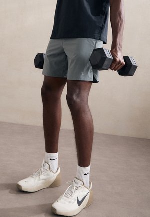 Pantaloncini atletici grigi con logo Nike, manubri esagonali neri e scarpe da ginnastica Nike beige con dettagli neri e calzini bianchi. Materiale testurizzato.