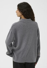 Woman with curly dark hair wearing a loose gray long-sleeve sweater with buttoned cuffs, viewed from the back against a plain background.