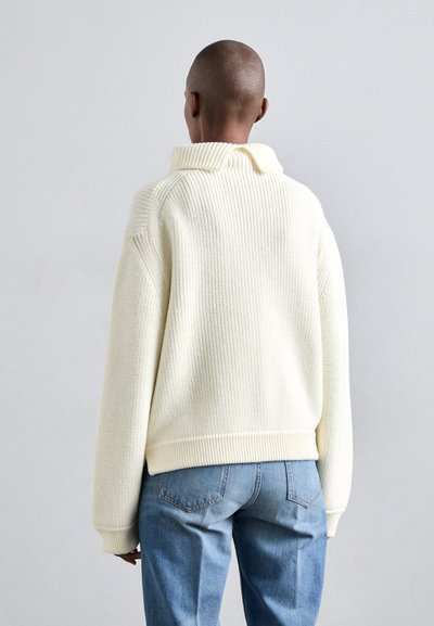 Pulover tricotat de culoare crem cu guler înalt, textură ribbed și umeri căzuți, asortat cu blugi de denim de culoare albastru deschis.