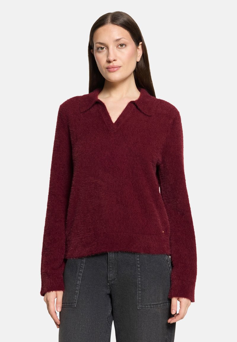 Femme debout portant un pull duveteux bordeaux à manches longues avec col et un pantalon gris foncé.