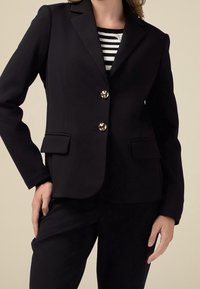 Blazer noir sur mesure avec un design structur é, deux poches frontales et des boutons dorés en accent. Porté par-dessus un haut rayé noir et blanc.