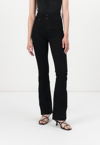 Femme portant un jean noir évasé taille haute et des sandales à talons noires à brides, debout devant un fond blanc uni.
