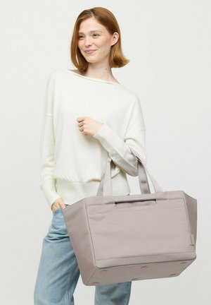 Vrouw met rood haar draagt een witte trui en lichtgekleurde spijkerbroek en houdt een grote beige boodschappentas met twee hengsels vast.