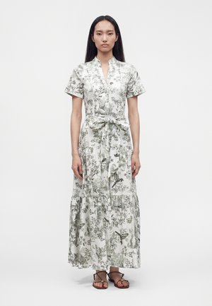 Femme aux longs cheveux foncés portant une robe longue blanche à motif floral, manches courtes, ceinture nouée à la taille et sandales marron, debout devant un fond uni.