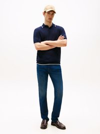 Camisa polo azul-marinho com detalhes brancos, calças de ganga azul e mocassins castanhos. O modelo usa um boné bege e está de braços cruzados em um fundo neutro.