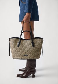 Borsa a spalla intrecciata con dettagli in pelle nera; presenta un motivo beige testurizzato, forma ampia e doppi manici. Abbinata con stivali alla caviglia marroni.