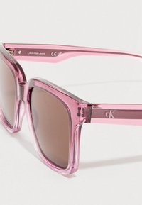 Óculos de sol de plástico rosa com armação quadrada, lentes castanhas tintadas e um logótipo na haste. Textura lisa com acabamento brilhante.