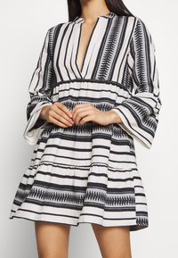 Robe rayée blanche et noire avec un col en V profond, des manches longues évasées et une jupe à volants. Présente des motifs géométriques et des accents texturés.