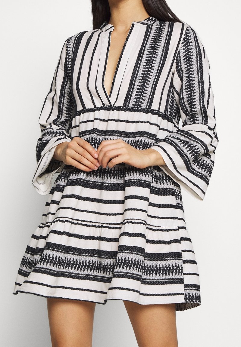 Robe rayée blanche et noire avec un col en V profond, des manches longues évasées et une jupe à volants. Présente des motifs géométriques et des accents texturés.