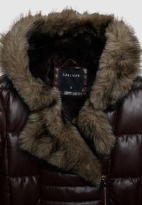 Giacca di pelle marrone scuro con un spesso colletto in pelliccia marrone e fodera interna, con etichetta del marchio Calliope visibile e una zip dorata.