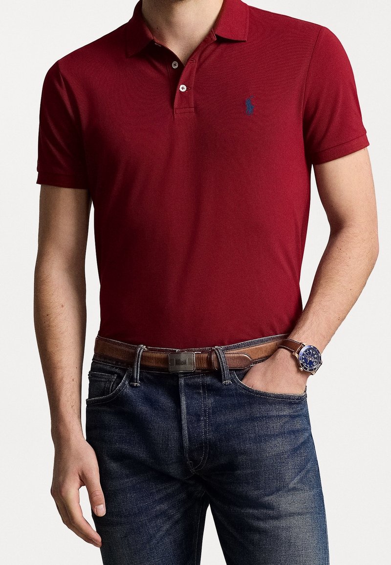 Polo rouge avec col, manches courtes et logo brodé bleu. Associé à un jean foncé et une ceinture marron. Les accessoires incluent une montre.