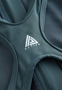 Gros plan du tissu d'un soutien-gorge de sport bleu sarcelle avec des coutures courbées et le logo blanc "ACTIVE" imprimé près du centre de la poitrine.