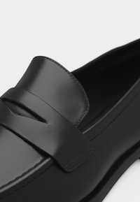 Mocassin en cuir noir avec une lanière élégante sur le dessus, une finition lisse et un design à bout arrondi.