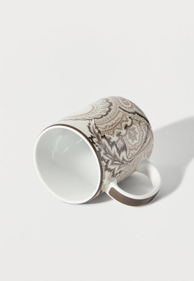 Tasse en céramique blanche ornée de motifs floraux et paisleys marron complexes, couchée sur le côté avec la poignée visible sur un fond blanc.