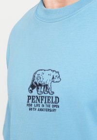 Jasnoniebieska koszula z czarnym haftowanym logo niedźwiedzia oraz napisem "Penfield For Life In The Open 50. rocznica" na klatce piersiowej.