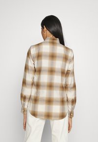 Pitkähihaisen ruudullisen paidan beige- ja ruskeasävyissä, jossa on kauluksellinen kaula-aukko, nappihaalari ja kaareva helma. Pehmeää puuvillakangasta.