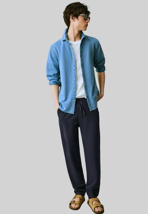 Camisa de botones azul claro, camiseta blanca, pantalones de chándal azul marino oscuro y sandalias marrones, llevados por una persona con cabello rizado y gafas.