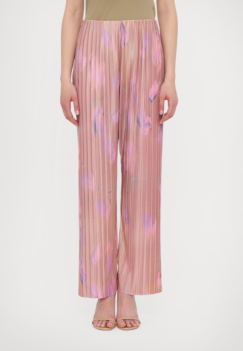 Femme portant un pantalon large plissé avec un motif floral rose et violet et des sandales à talons beiges, debout sur fond blanc.