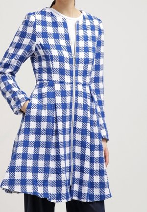 Manteau à manches longues avec motif à carreaux bleu et blanc, fermeture éclair devant et silhouette évasée, porté sur un haut blanc et un pantalon foncé.