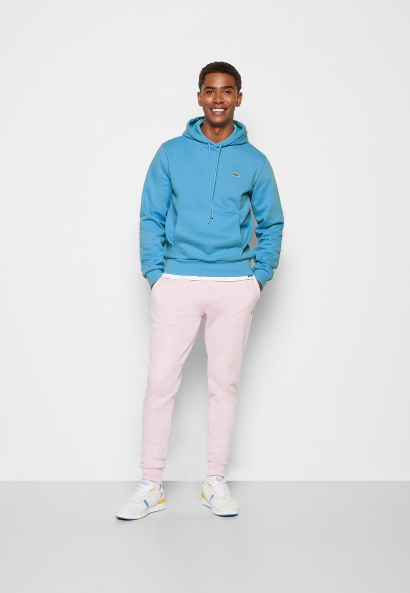 Lichtblauwe hoodie met een voorzak, gecombineerd met zachte pastelroze trainingsbroek en witte sneakers met gele en blauwe accenten.
