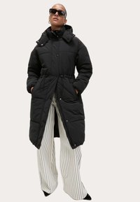 Chaqueta exterior acolchada negra con cintura ceñida, cuello alto y cierres de botón. Combinada con pantalones de pierna ancha de rayas blancas y zapatos negros.