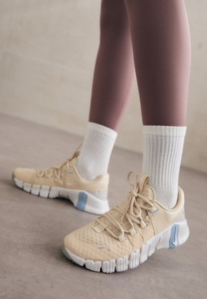 Person iført beige og hvide atletiksneakers med hvide ankelsokker, stående på et glat indendørs gulv.