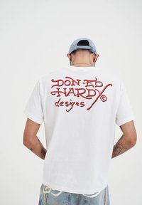 Bílý bavlněný tričko s červeně vyšitým nápisem na zádech, který zní "DON ED HARDY designs." Krátké rukávy a uvolněný střih.
