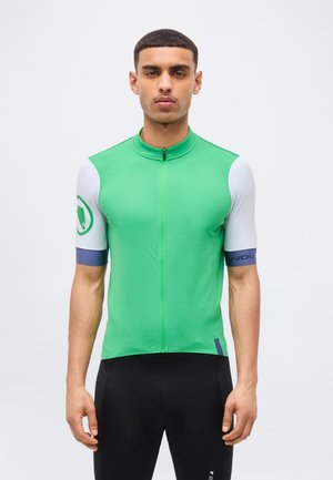 Νέος άντρας που φοράει μια πράσινη και λευκή ποδηλατική φανέλα με κοντά μανίκια, φερμουάρ στο μπροστινό μέρος και μαύρο ποδηλατικό σορτς, στέκεται μπροστά σε λευκό φόντο.