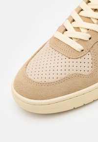 Baskets beige avec dessus en daim, accents perforés pour la respirabilité, lacets crème et semelle en caoutchouc texturée. Finition lisse et douce.