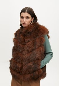 Umělá kožešinová vesta v bohatých hnědých tónech, s vzorem ve tvaru písmene V, silnou texturou a vysokým límcem. V kombinaci se zeleným topem s dlouhým rukávem.