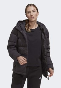 Black puffer jacket with a hood, featuring a shiny, quilted texture and snap button closures. Worn over a black t-shirt and sweatpants.